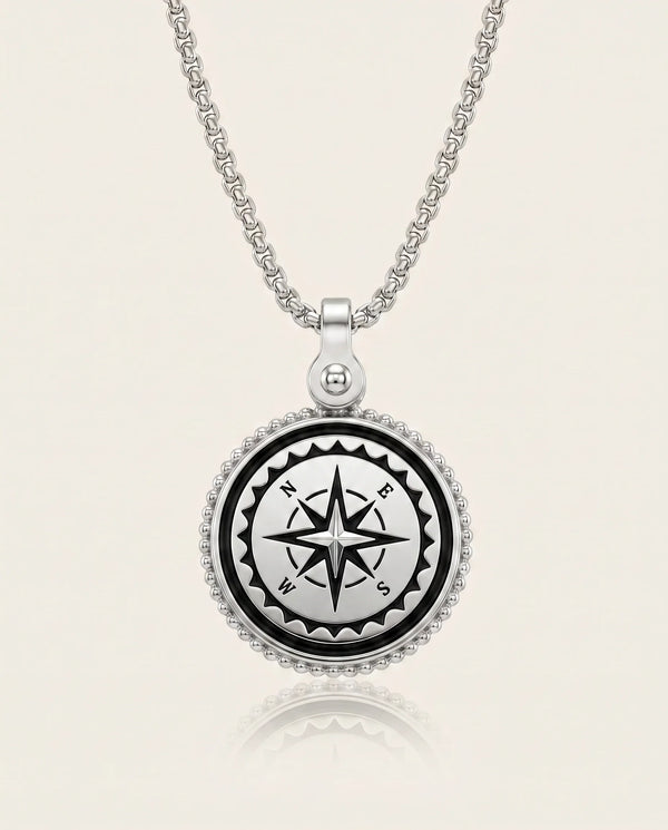A silver compass pendant necklace on a matching chain, featuring a black compass rose design in the center with white lettering, surrounded by a beaded silver border, set against a light cream background.