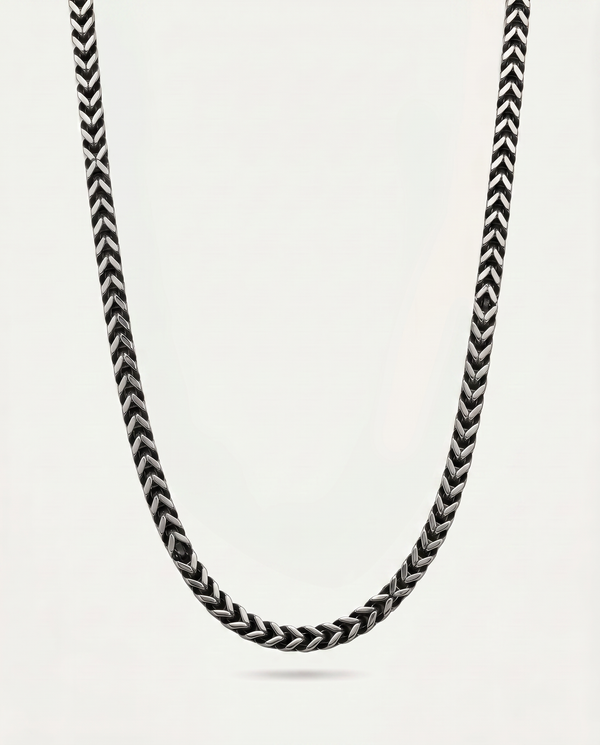 A close-up of a braided chain necklace in black and white, suspended against a plain light gray background.