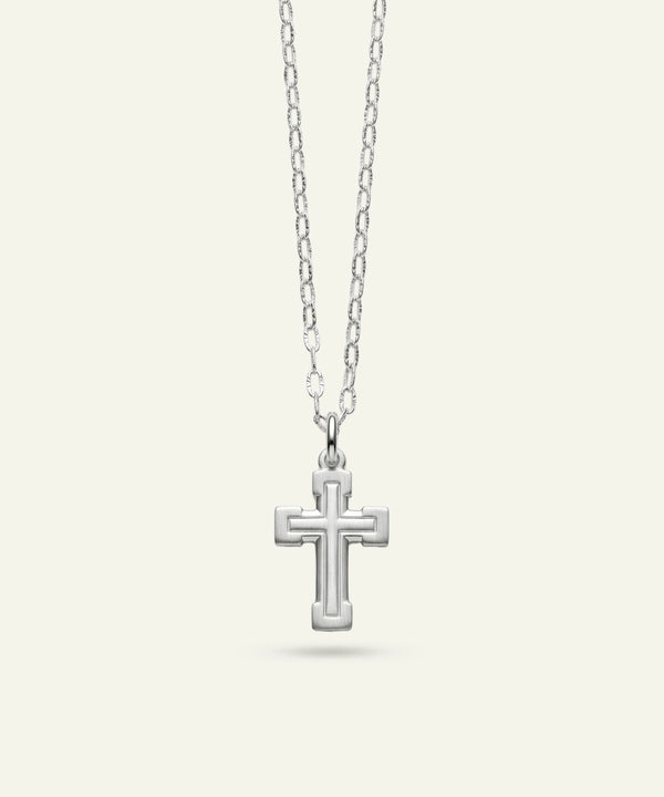 A silver cross pendant necklace with a textured, raised geometric border design, suspended from a delicate silver link chain against a light cream background.