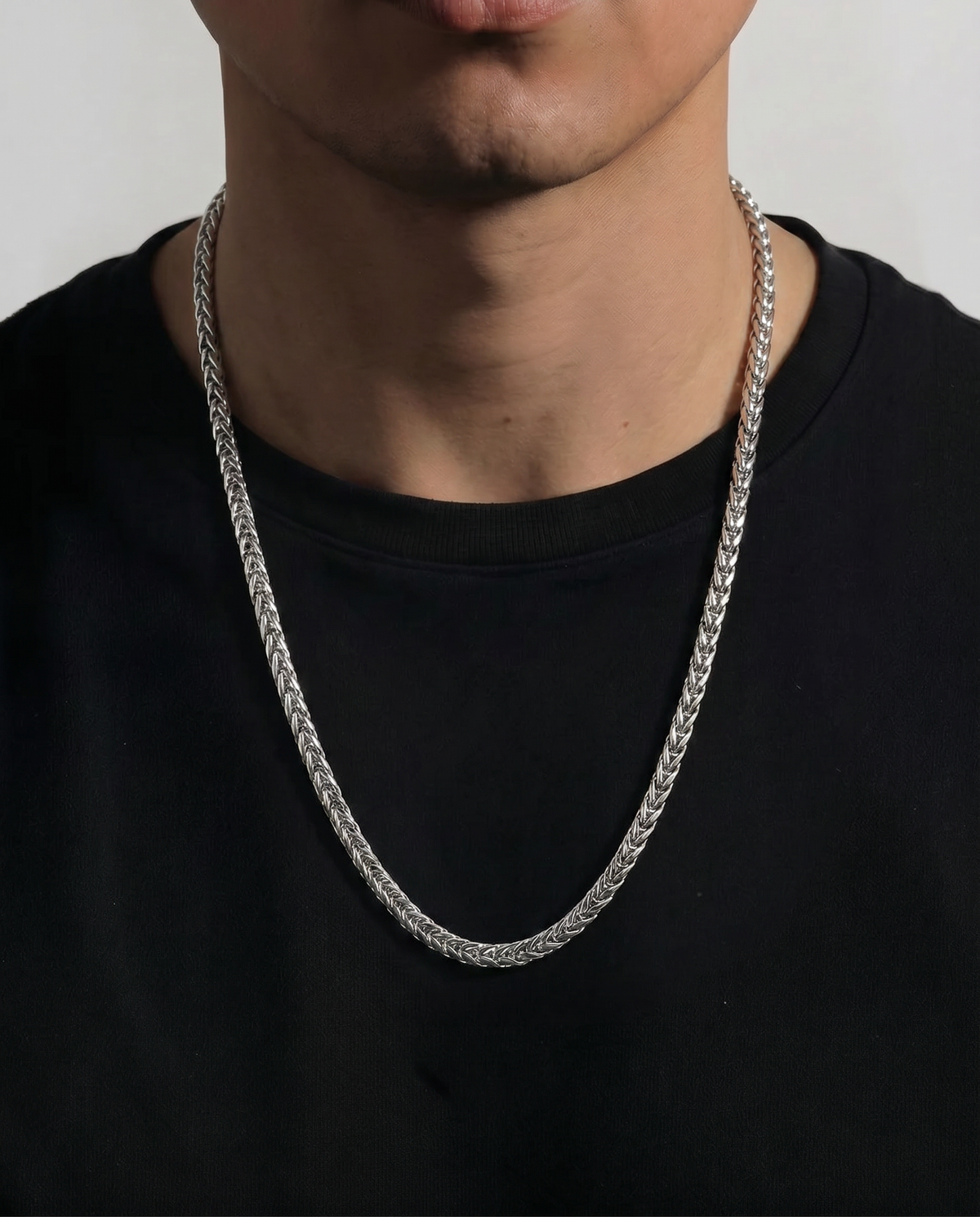 A close-up of a person wearing a thick silver chain necklace over a black t-shirt against a light gray background.