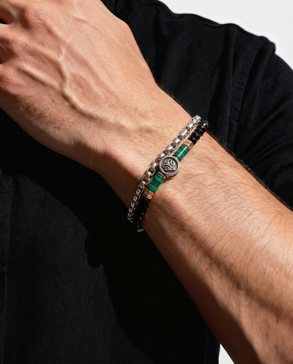A close-up of a man's forearm wearing a stack of three bracelets. The bracelets include a silver chain with clear stones, a black beaded bracelet with green accents, and a silver chain with a circular medallion.