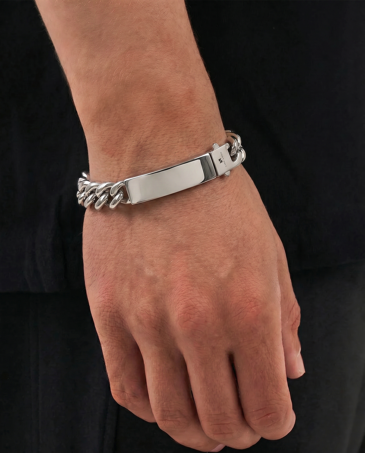 A silver-toned metal bracelet with a polished rectangular plate and curb-link details, shown worn on a person's wrist against a black shirt background.