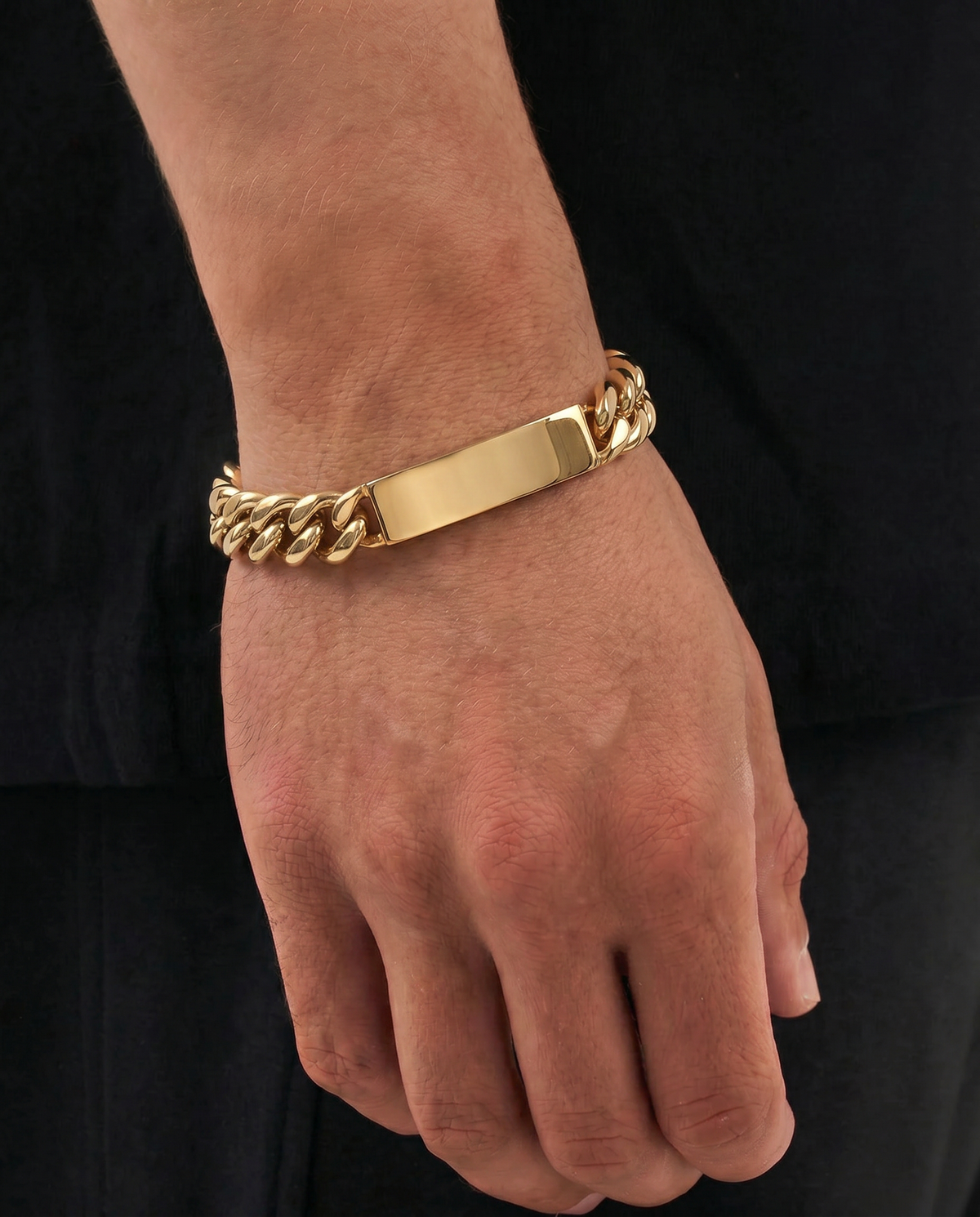 A gold-toned metal ID bracelet with a flat rectangular plate, worn on a person's wrist against a black shirt background.