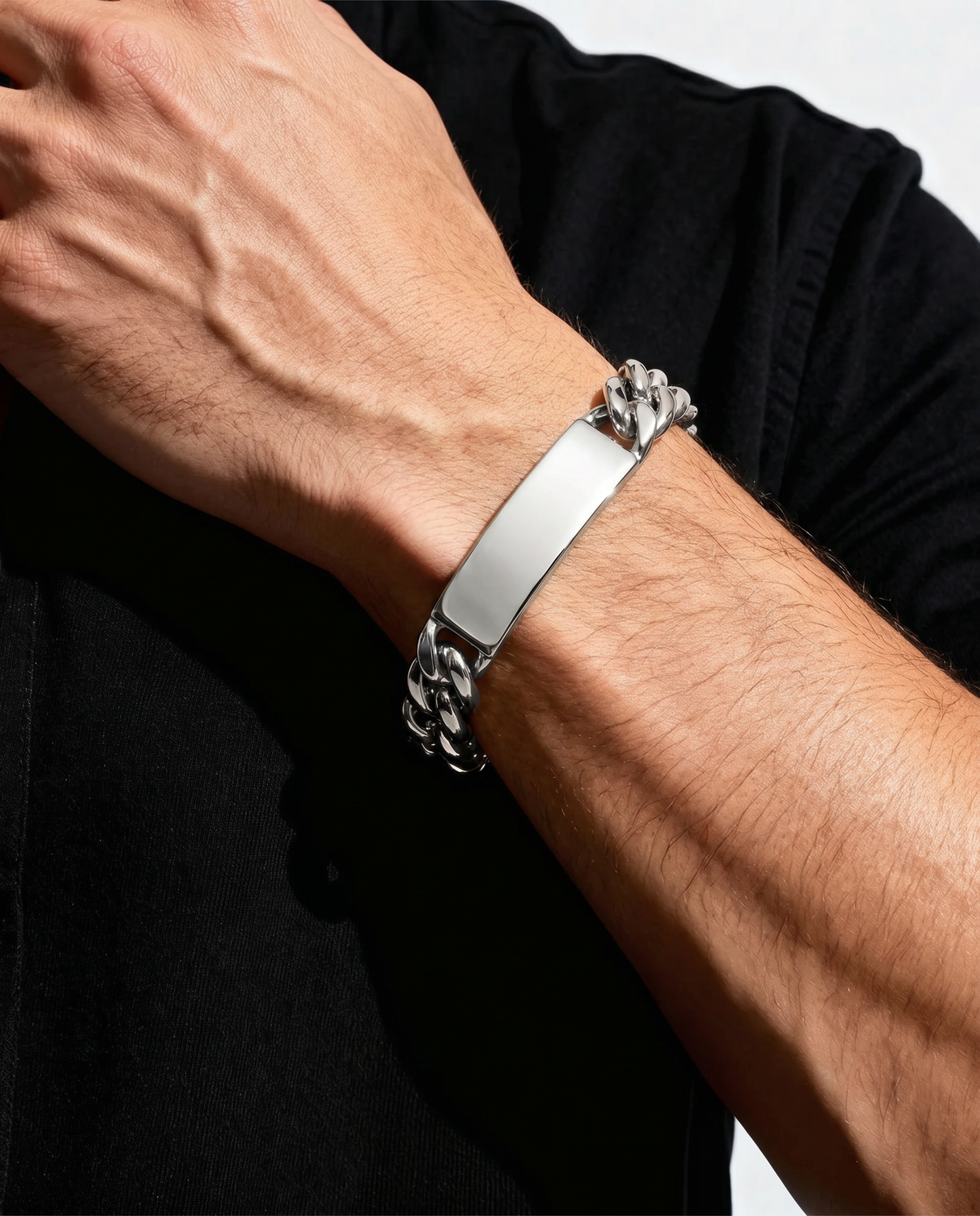A close-up of a man's forearm wearing a silver-toned metal bracelet with a polished rectangular plate and a textured chain-link design on the ends, worn over a black shirt.
