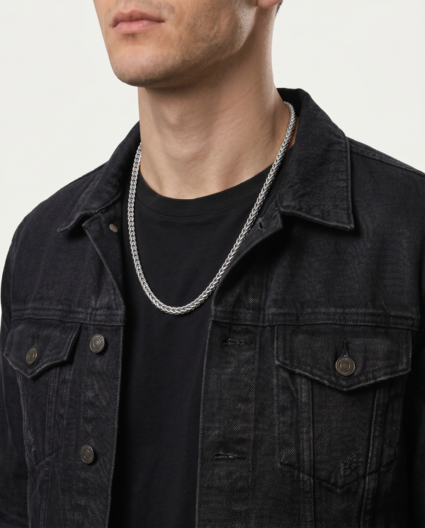 A close-up of a man wearing a silver chain necklace over a black t-shirt and a dark washed denim jacket.