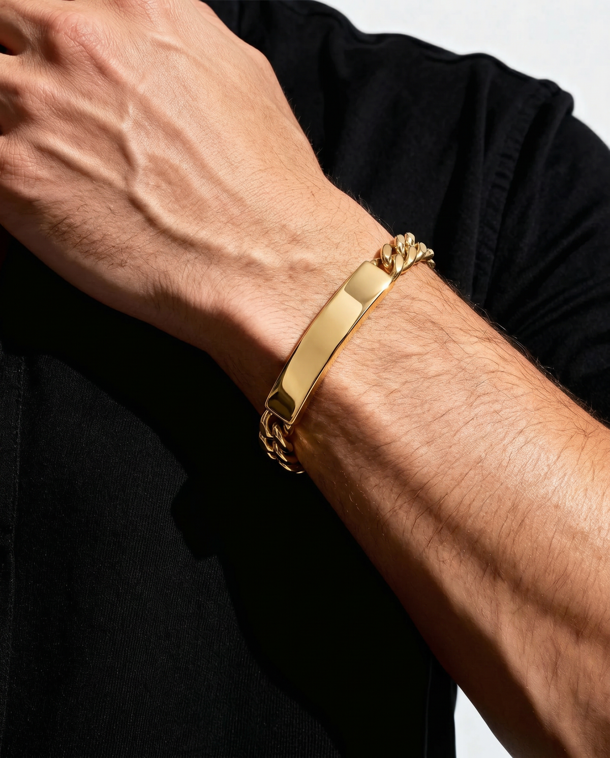 A close-up of a person's forearm wearing a gold-plated ID bracelet with a flat rectangular plate and a curb link chain detail, paired with a black shirt.