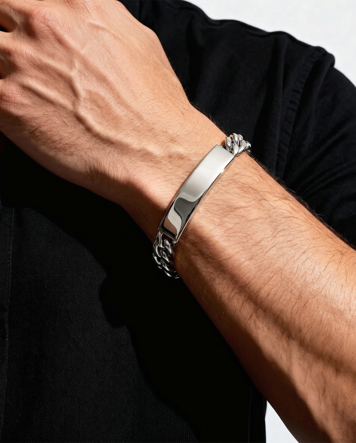 A close-up of a man's forearm and hand wearing a polished silver-tone metal cuff bracelet with a curb chain detail on one end, set against a black shirt and light background.