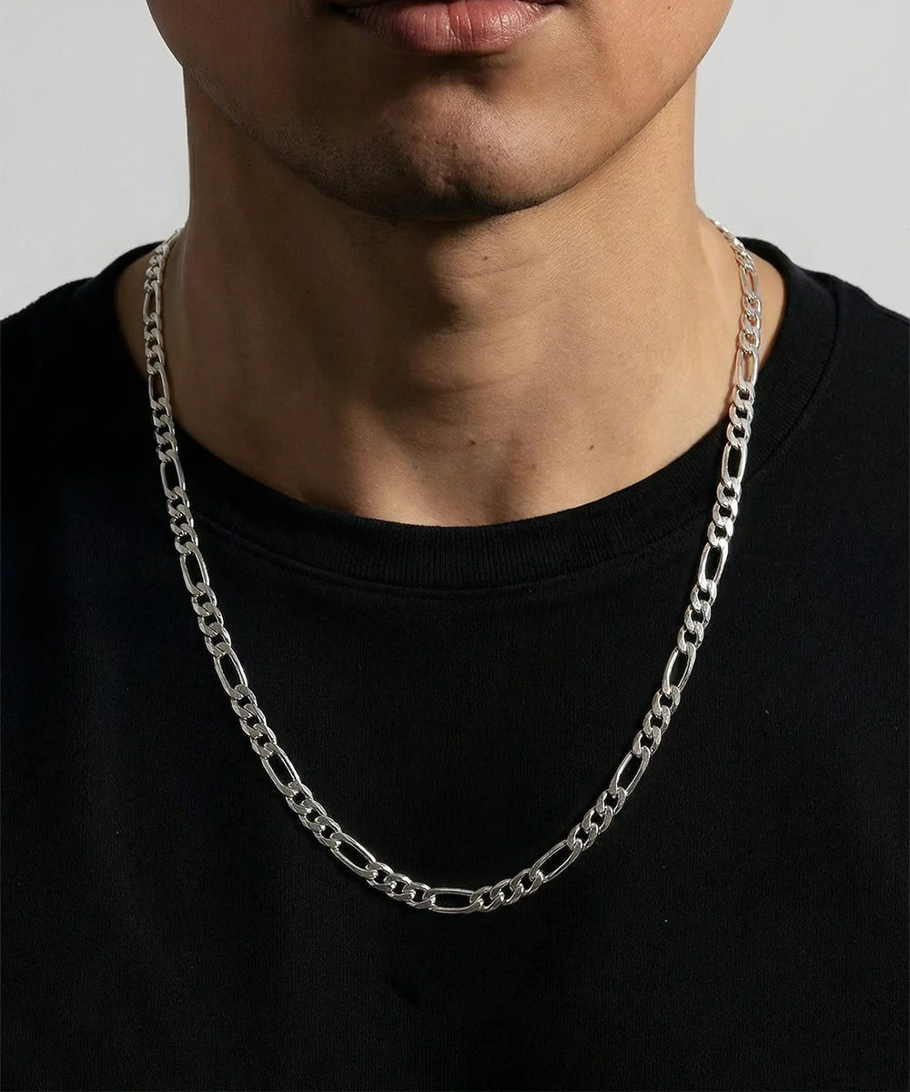 A close-up of a person wearing a silver Figaro chain necklace over a black crewneck t-shirt, cropped from the chin down.