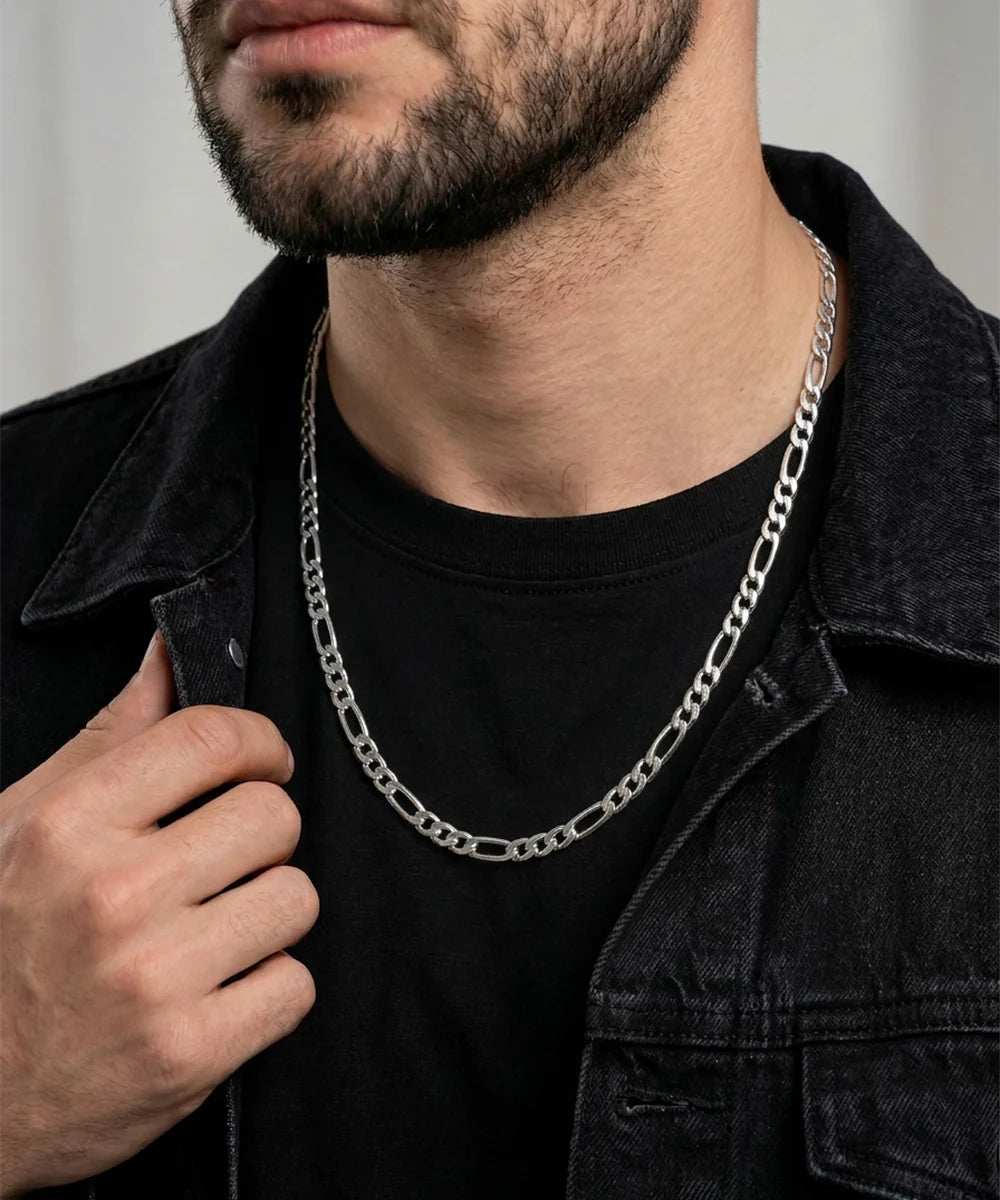 A close-up of a man wearing a silver Figaro chain necklace over a black t-shirt and a dark denim jacket.