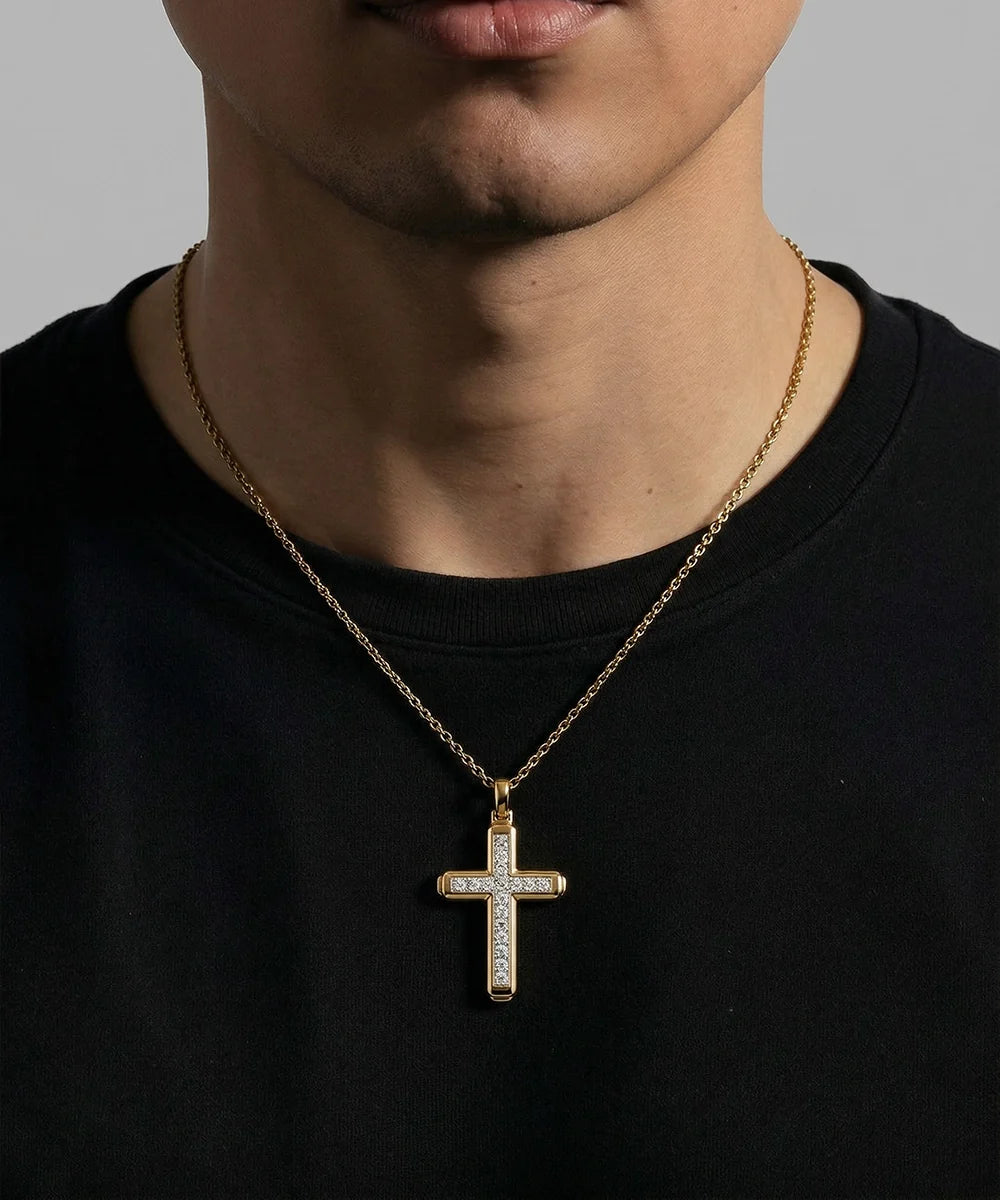 A gold cross pendant necklace with clear stones, worn by a model in a black t-shirt against a neutral gray background.