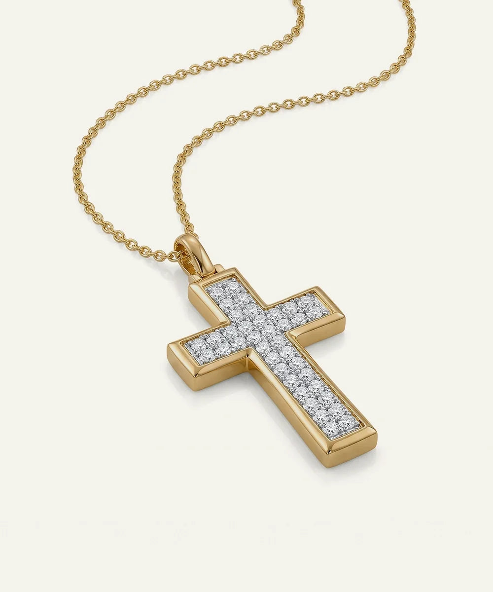 A gold cross pendant necklace paved with small clear stones, shown on a delicate gold chain against a light cream background.