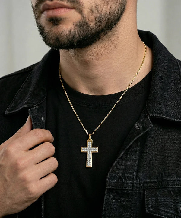 A close-up of a man wearing a gold cross pendant necklace with clear stones, styled over a black t-shirt and a dark denim jacket.