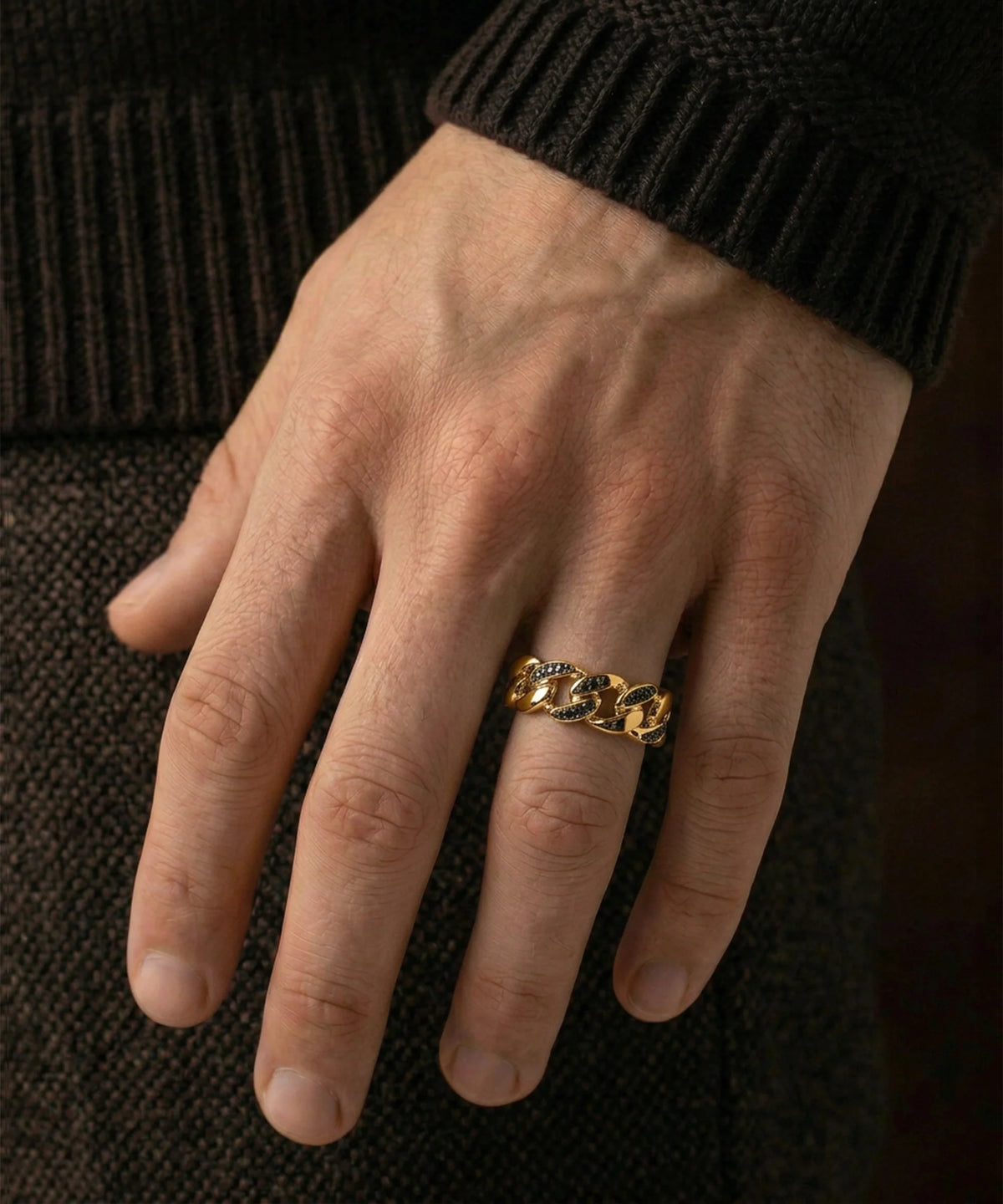 A person's hand wearing a men's yellow gold Cuban link ring set with black stones against a dark background.