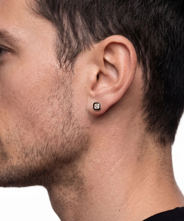 Close-up lifestyle shot of a man's side profile wearing a square gold stud earring featuring a dark central stone framed by a black halo border.