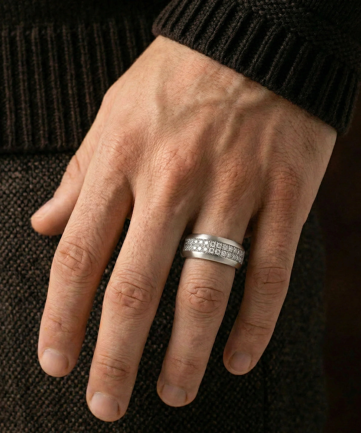 A person's hand wearing a men's silver two-row diamond band ring against a dark background.