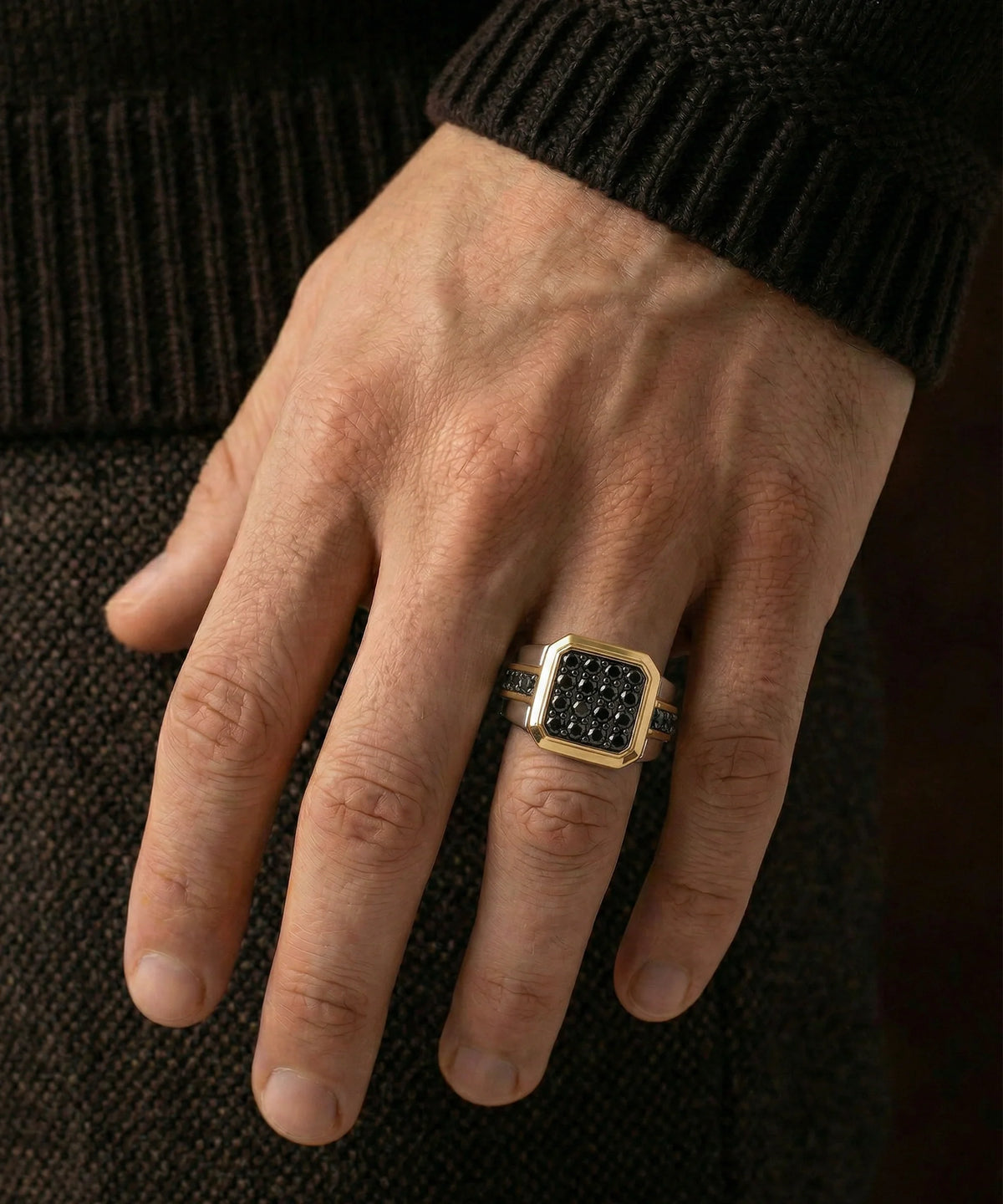 Close-up shot of a man's hands wearing a two-tone silver and gold composite ring featuring a cushion-shaped face paved with black diamonds and accent side rows, set against a dark background.