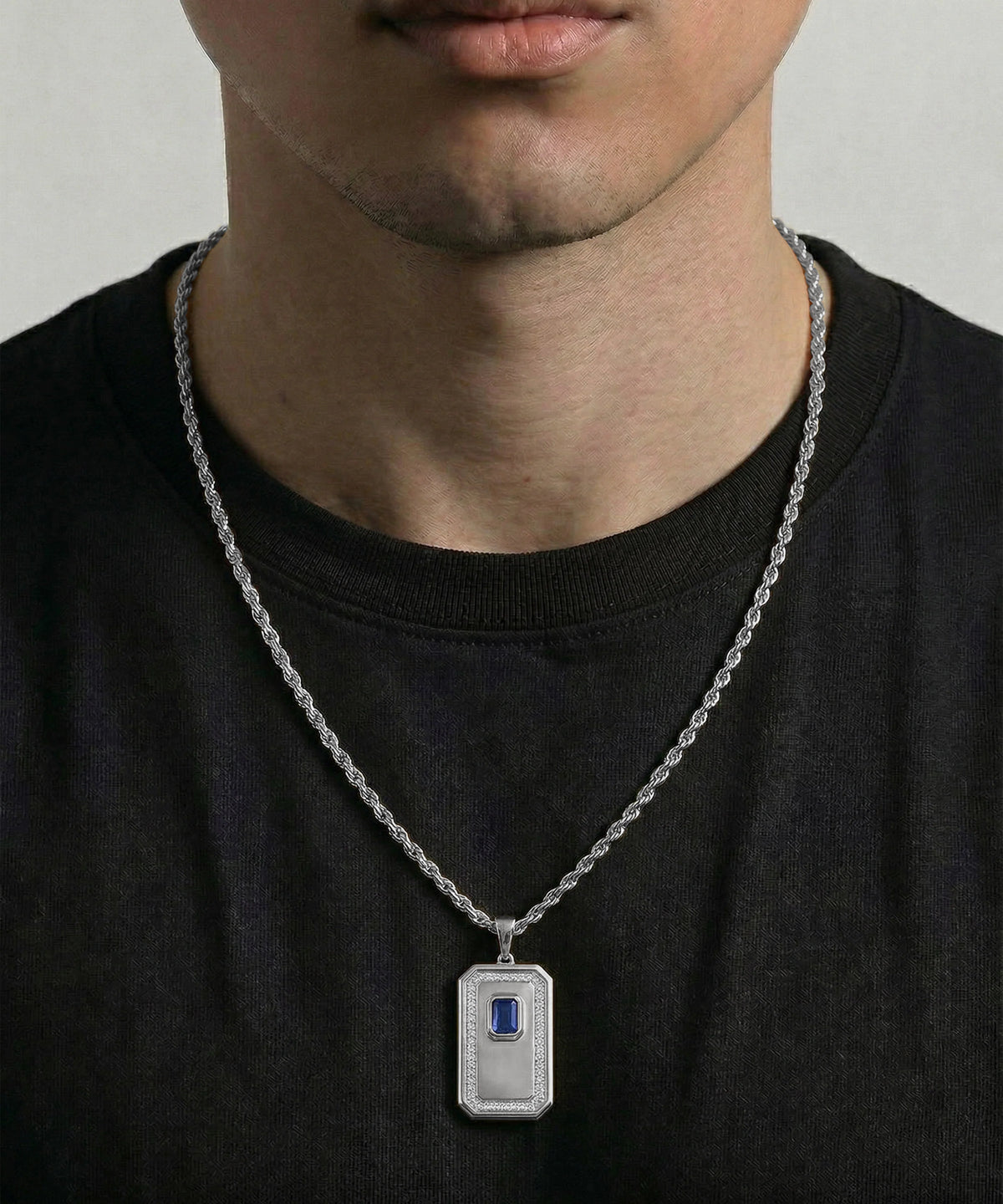 A close-up of a man wearing a silver-toned metal pendant necklace with a rectangular badge-shaped charm featuring a small blue rectangular inset, paired with a black t-shirt.