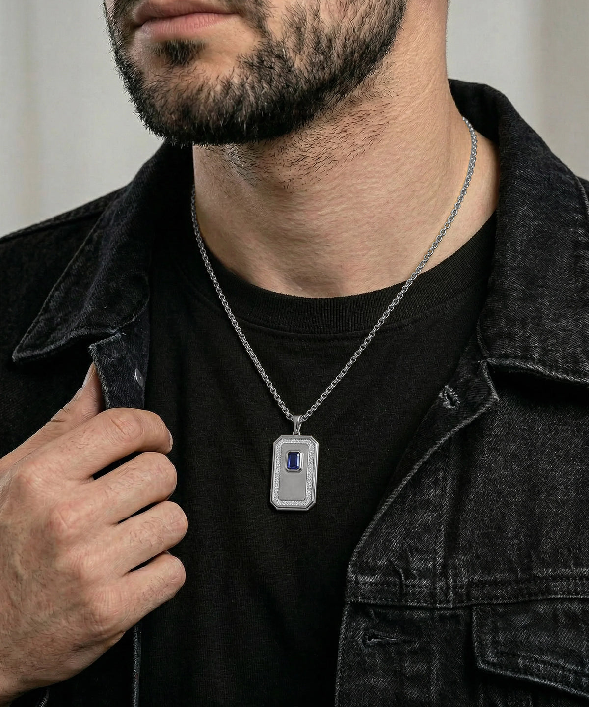 A close-up of a man wearing a silver-toned rectangular pendant necklace with a blue inlay, styled over a black t-shirt and dark denim jacket.