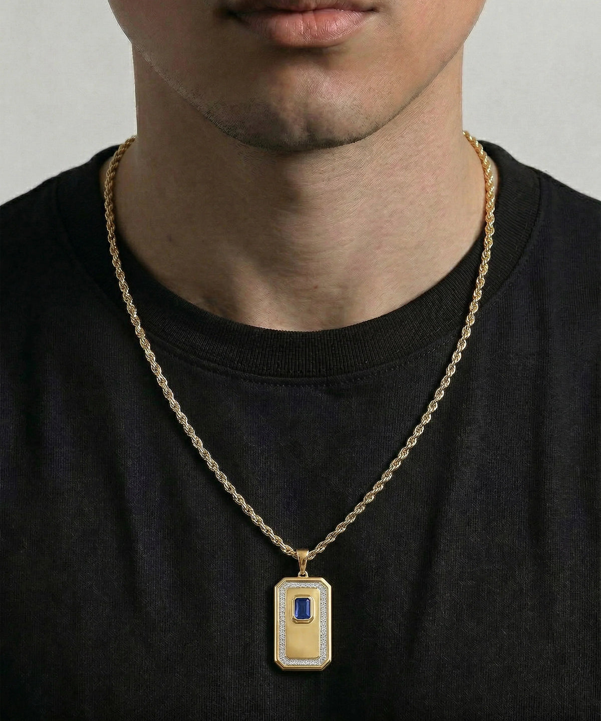 A close-up of a man wearing a gold chain necklace with a rectangular pendant featuring a yellow center, a blue rectangular inset, and a white textured border.