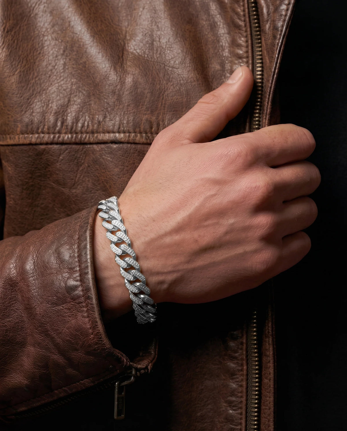 Close-up lifestyle shot of a man's wrist wearing a silver-toned pavé diamond-encrusted Miami Cuban chain bracelet, styled against a brown leather jacket.