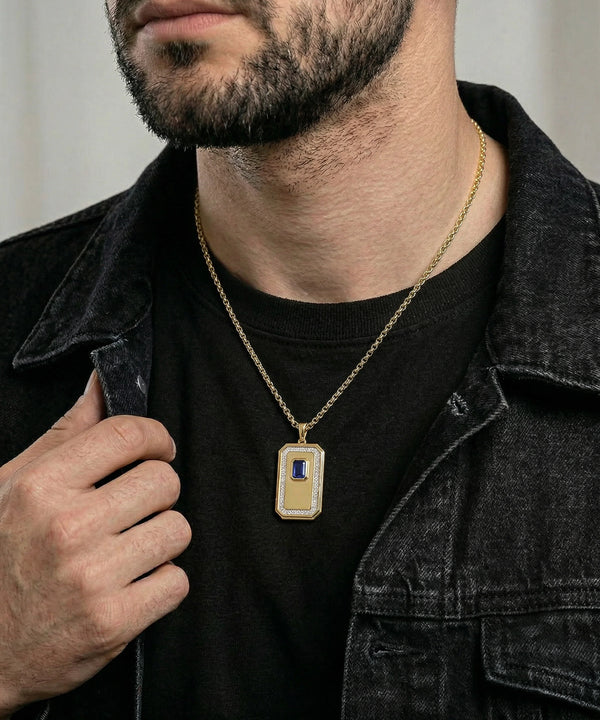 A close-up of a man wearing a gold rectangular pendant necklace with a blue and white inlay, styled over a black t-shirt and dark denim jacket.