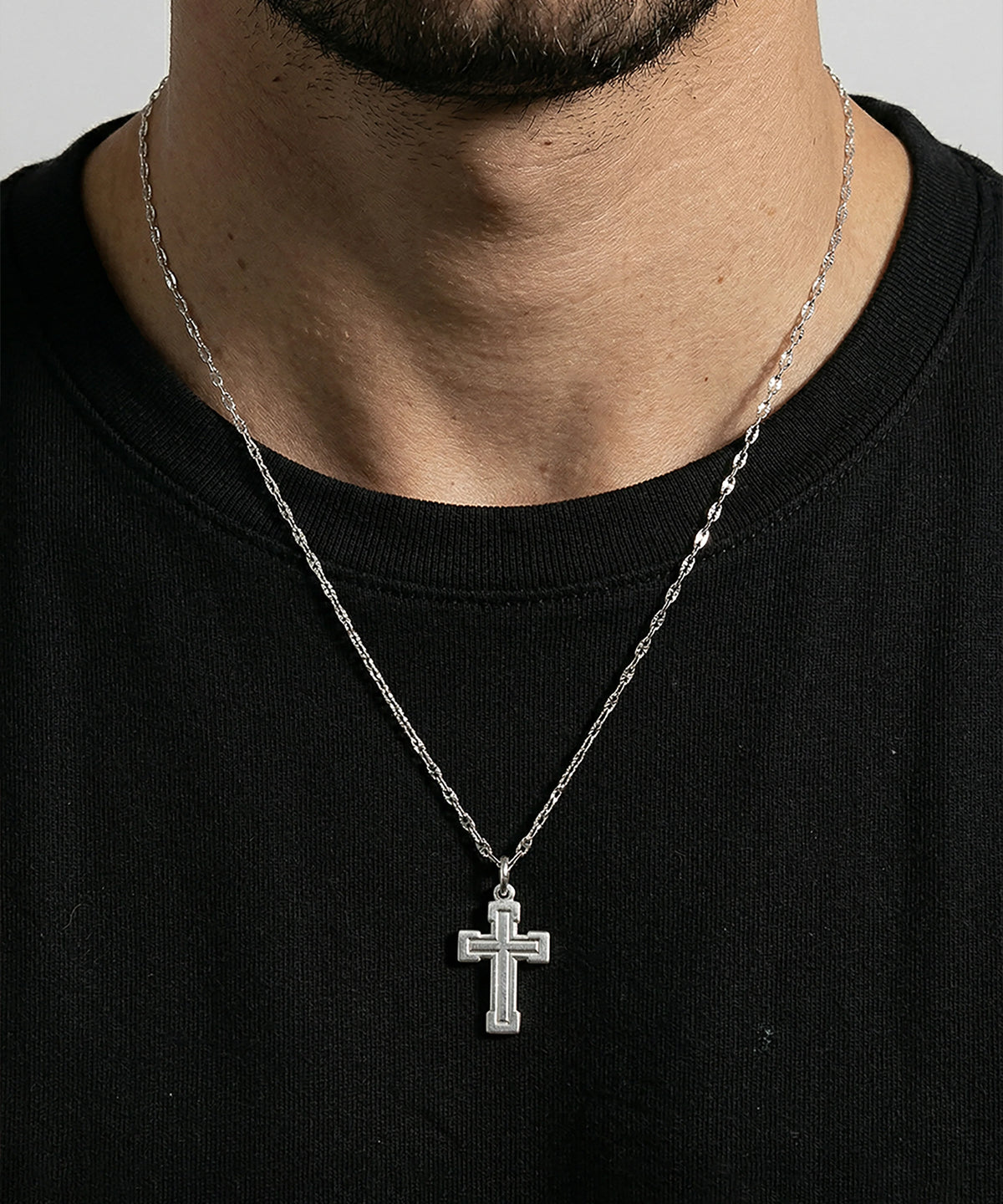 Close-up lifestyle shot of a man wearing a silver cross pendant necklace with a textured border design, suspended from a thin link chain over a black t-shirt