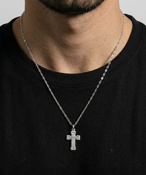 Close-up lifestyle shot of a man wearing a silver cross pendant necklace with a textured border design, suspended from a thin link chain over a black t-shirt