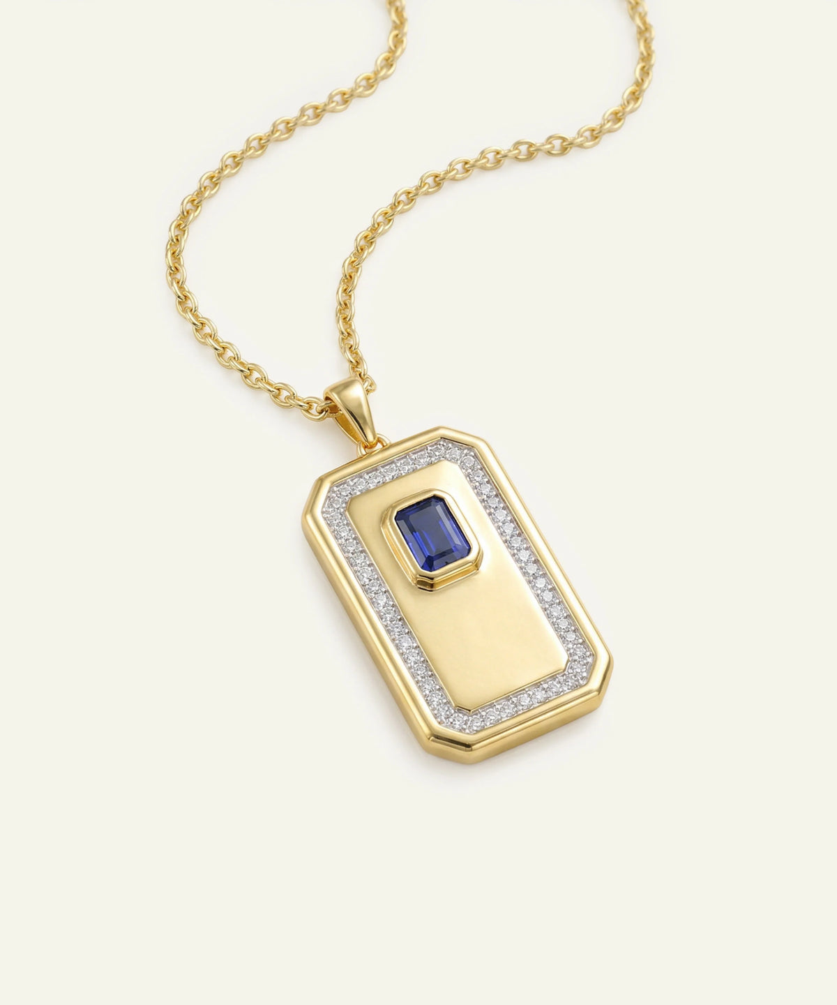 A gold pendant necklace featuring a rectangular dog-tag-style pendant with a blue emerald-cut gemstone in the center, surrounded by a border of small clear stones, shown on a light cream background.