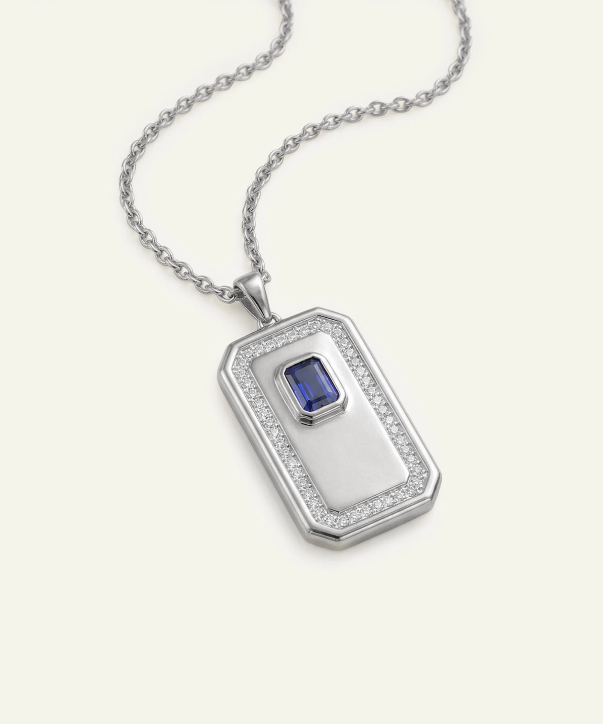 A silver-toned rectangular pendant necklace featuring a central blue emerald-cut gemstone surrounded by a halo of small clear stones, suspended from a delicate chain on a light cream background.